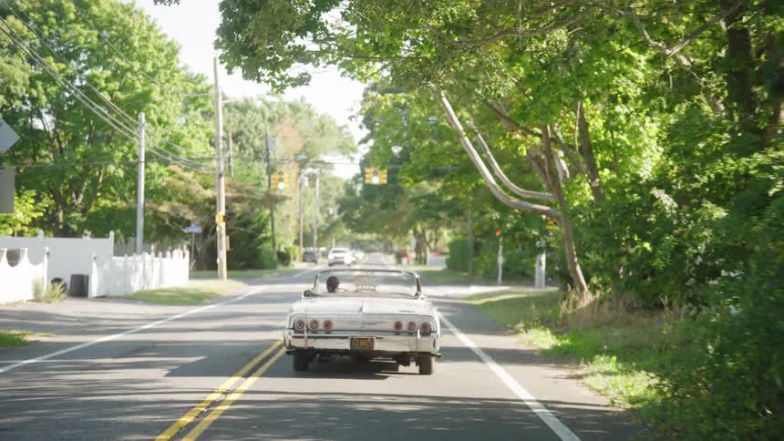 The video is a stationary medium shot of a white vintage convertible driving away down a treelined suburban road on a sunny day the road is paved bordered by green foliage and white picket fences and the classic car has its top down