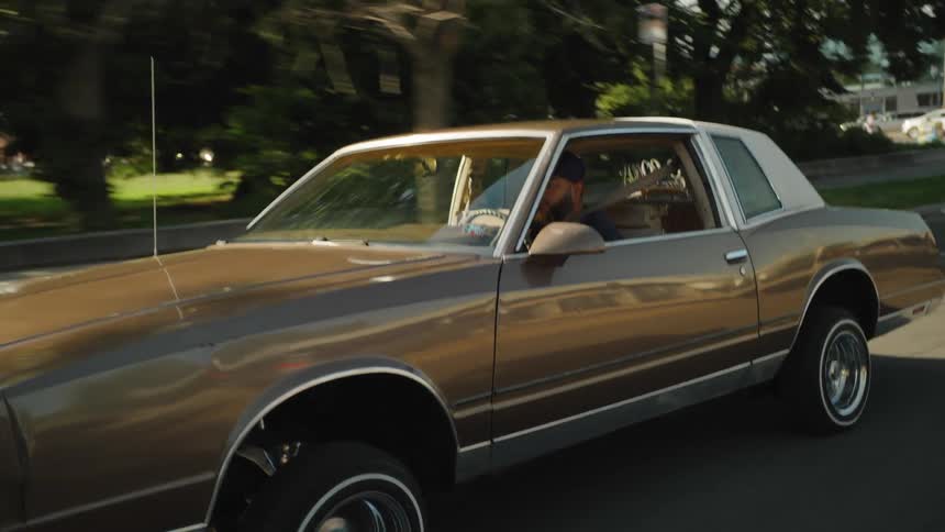 A young man with a long beard drives a brown lowrider down the street on a sunny day the car bouncing as it moves