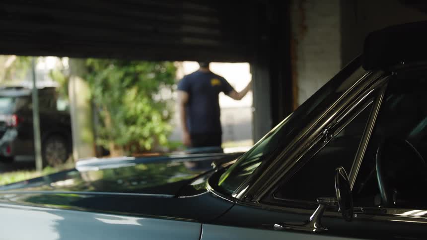 A dark garage door slowly rolls upward revealing the bright outside world the front section of a classic car sits in the dark foreground and a man stands in the doorway looking inward after the door is fully raised