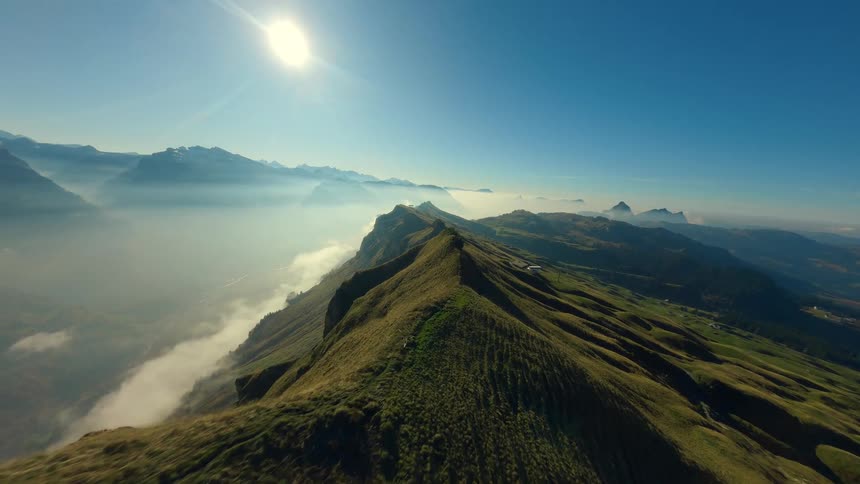 A fast sweeping aerial shot flies over a green mountain ridge with steep cliffs overlooking a valley filled with low hanging fog and snow capped peaks in the distance under a bright sunny sky