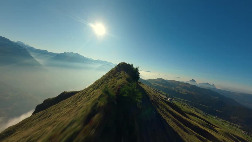 An aerial shot speeds towards a person on a sharp mountain peak zips past them and soars rapidly along the grassy ridgeline before slowing to reveal the vast cloudfilled valleys