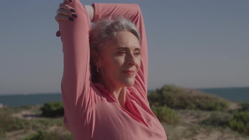 A woman with gray hair performing an arm stretch with her hands clasped behind her head standing on vegetated dunes near the ocean under a clear sky