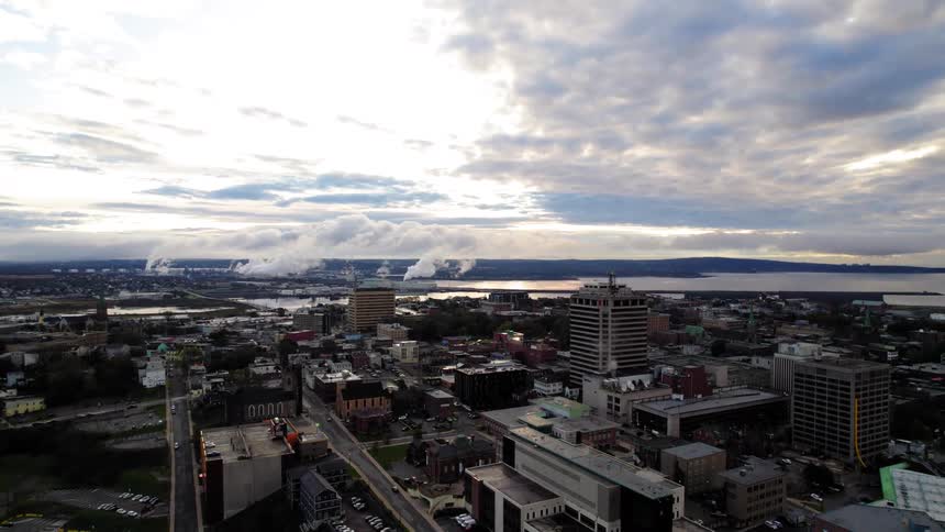 An aerial view shows an urban city center with highrise buildings and lower structures a large harbor is visible in the background thick plumes of steam rise from an industrial area near the water and the sky is covered with scattered clouds