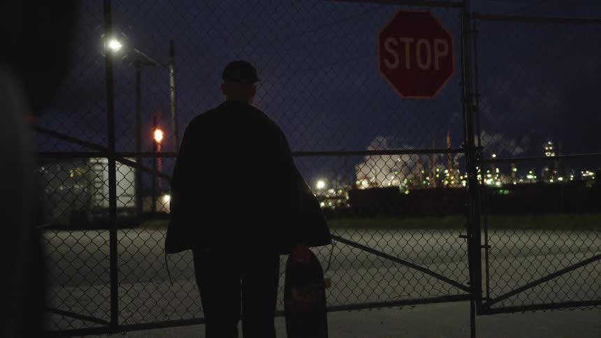 The video shows a silhouetted person wearing a cap and holding a skateboard standing in front of a chainlink fence gate at night a prominent stop sign is affixed to the gate and in the background a brightly lit industrial facili