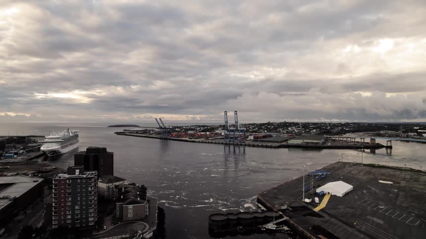 An aerial extreme wide view shows a cruise ship docked next to a busy industrial port with blue cargo cranes surrounded by urban buildings and dark choppy water under an overcast sky