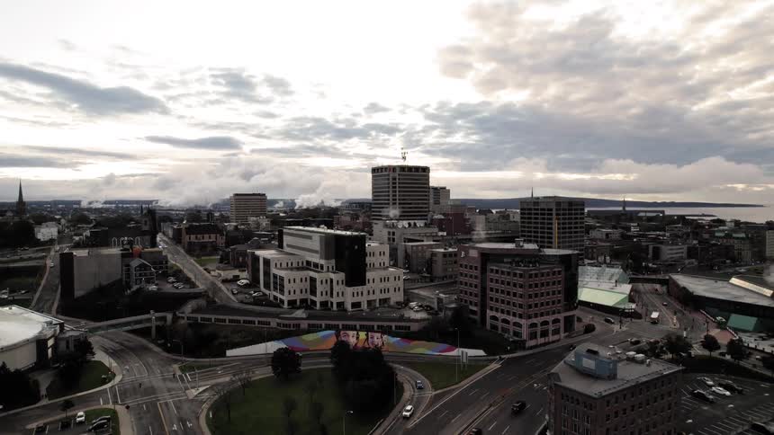 An aerial view shows a compact city skyline dominated by midrise office buildings and a road network in the foreground all under a dramatic clouded sky with a body of water in the distance
