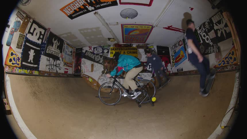 The video is shot with a fisheye lens and shows a group of people socializing and doing extreme sports in a small indoor skate parkhalfpipeone person is skateboarding across the curve while another is riding a bicycle on the flat bottom