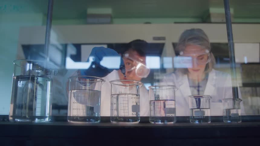 A line of glass beakers filled with clear liquid behind a glass shield while two people wearing lab coats and eye protection work on an experiment with one person adding a dark liquid that causes a color change in one of the beakers