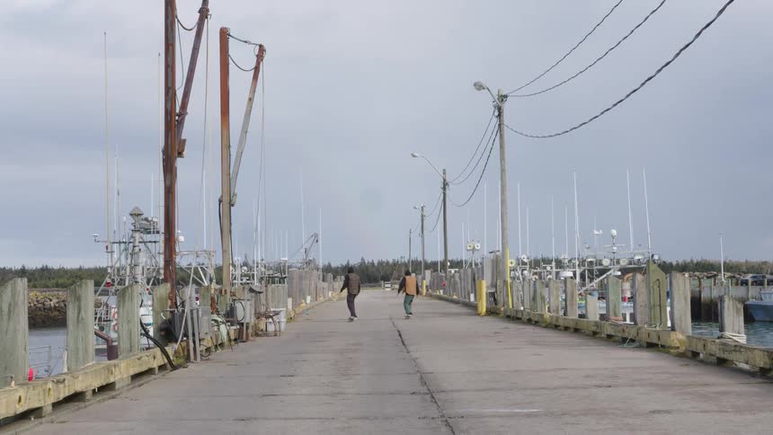 Two skateboarders ride along a pier surrounded by fishing boats