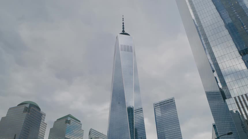 The video is an extreme lowangle shot focusing on the towering height of the one world trade center skyscraper which dominates the frame and points toward a grey overcast sky it is surrounded by several other modern glass office buildings