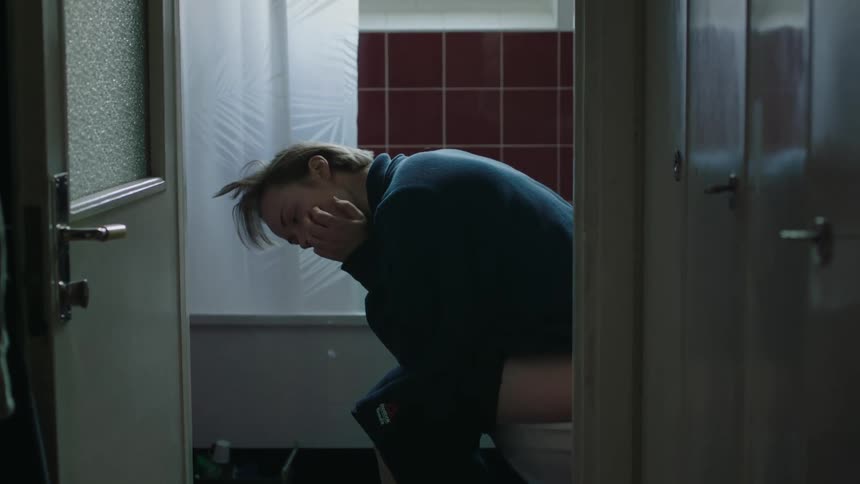 A young woman sits hunched over on a toilet in a brightly lit bathroom covering her face with the hand in distress and the scene is framed by the doorway and a dark hallway
