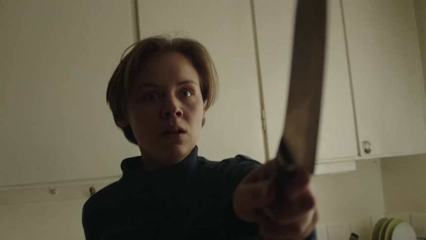 A young woman with short hair turns quickly in an indoor kitchen holding a large knife out toward the camera with a shocked expression on their face and white kitchen cabinets and a countertop are visible