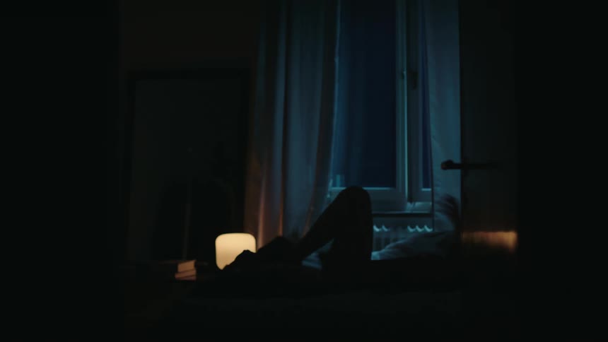 A silhouette of a person getting into bed turning off a glowing table lamp and sitting up near a window with blue ambient light in a dark bedroom