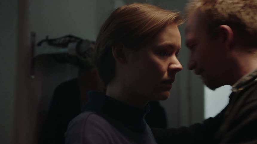 A man and a woman emerge through a doorway from a brightly lit kitchen into a darker hallway where the woman turns to the side and the man embraces her followed by the woman standing alone with a somber expression