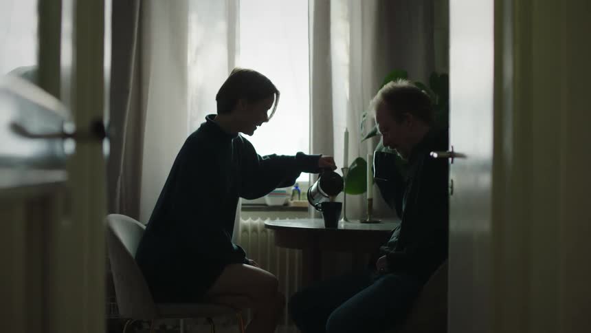 The video shows an interior shot of two people sitting at a table in a room with a bright window framed by the dark foreground of a doorway and a chair a younger person silhouetted against the light pours a drink from a french press coffee maker f