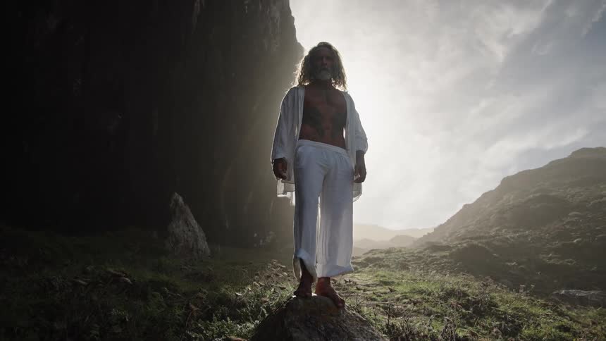 Longhaired man in white clothing standing on a rock in a sunlit valley