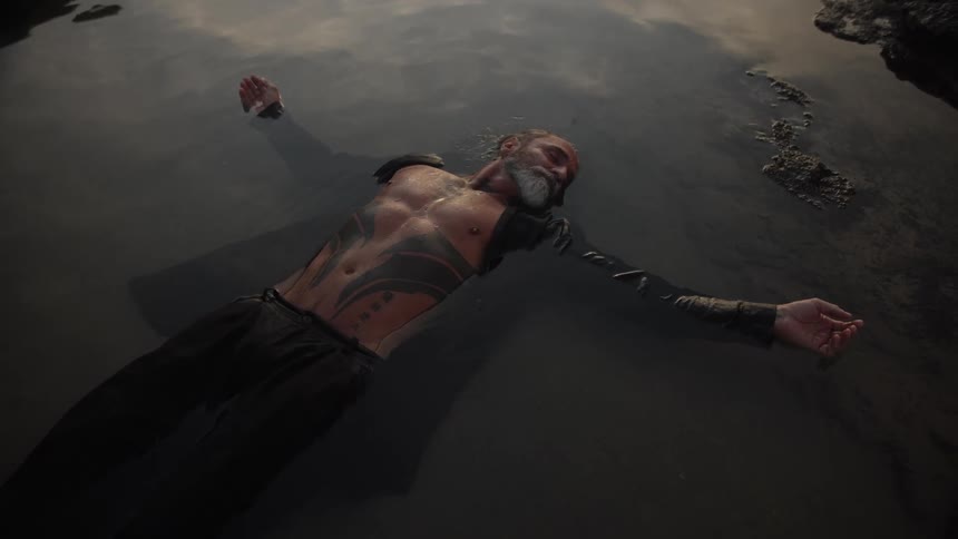A medium high angle view shows a bearded and tattooed man with long wet hair floating on his back in dark calm water near a rocky edge with soft daylight above