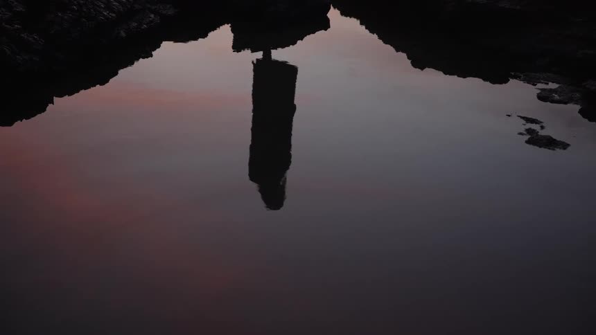 Reflection of upright figure on still water surrounded by rocky edges at dusk