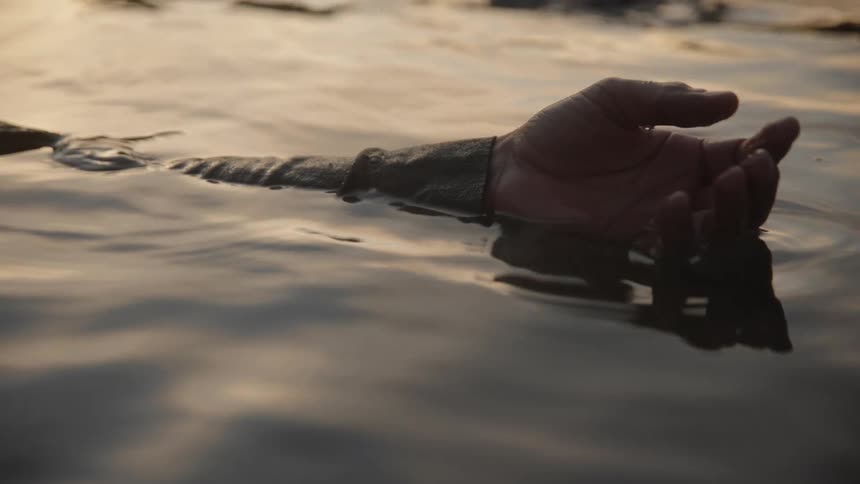 A close view of a dark hand and forearm resting just above the surface of calm dark water with a bright sunset reflecting softly on the ripples