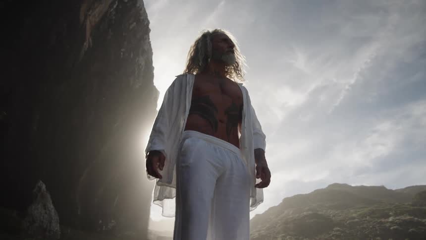 Man with long hair and beard lifts face toward light among rocky cliffs under cloudy sky