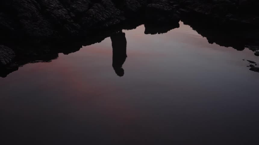 Reflection of person standing over still water surrounded by dark rocky terrain