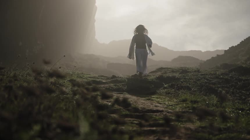 A person walks along a path between areas of rock and ground cover moving toward bright light beyond the surrounding cliffs and hills