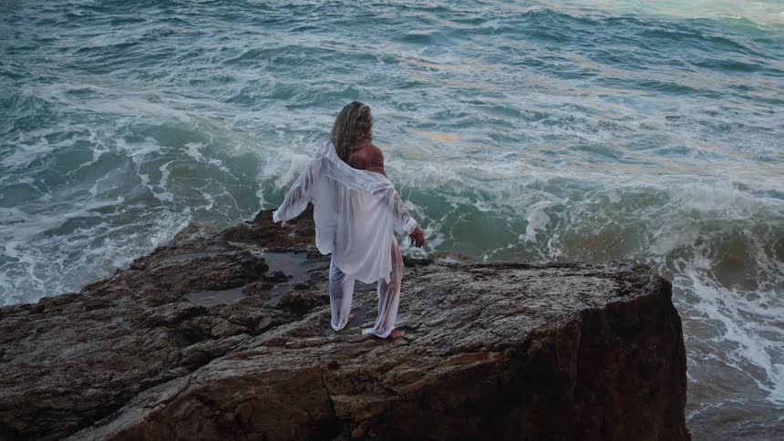 A wide high angle view shows a blonde man wearing a sheer white shirt and pants standing on a dark rocky cliff overlooking the ocean with his arms extended as waves crash below