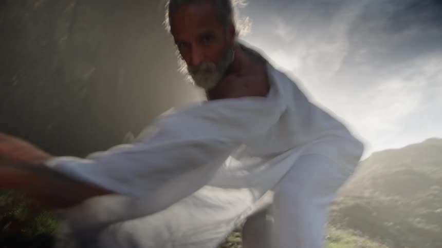 A man with a long beard moves vigorously in a dark misty setting swinging a large white piece of cloth with his outstretched arms toward the bright light source behind him