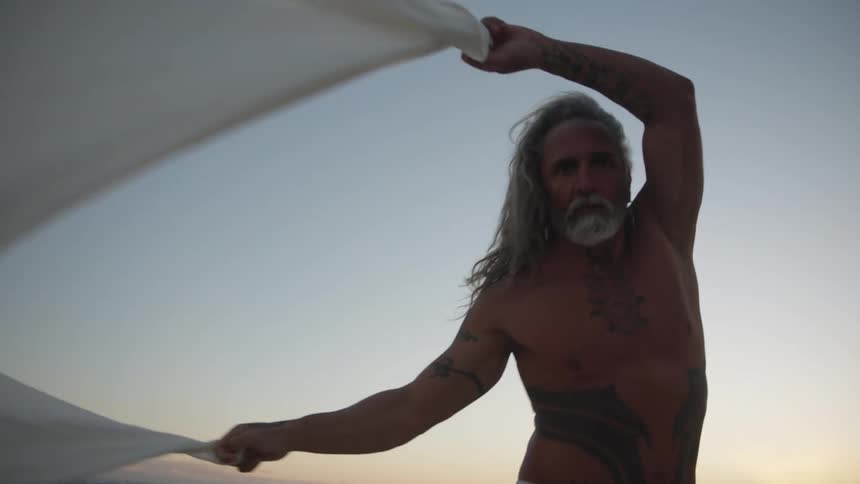 A close low angle view shows a shirtless older man with a gray beard and long hair wearing white pants and large black chest and arm tattoos holding a long piece of white cloth above his head on a beach at sunset
