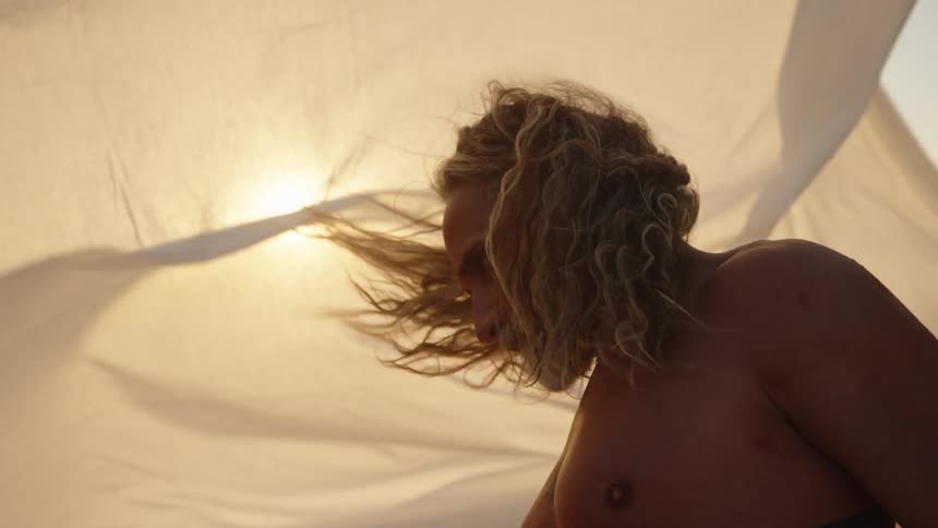 A close view shows a shirtless tanned man with long blonde curly hair and a gray beard standing against a white sunlit sheet with his arms raised over his head revealing arm tattoos