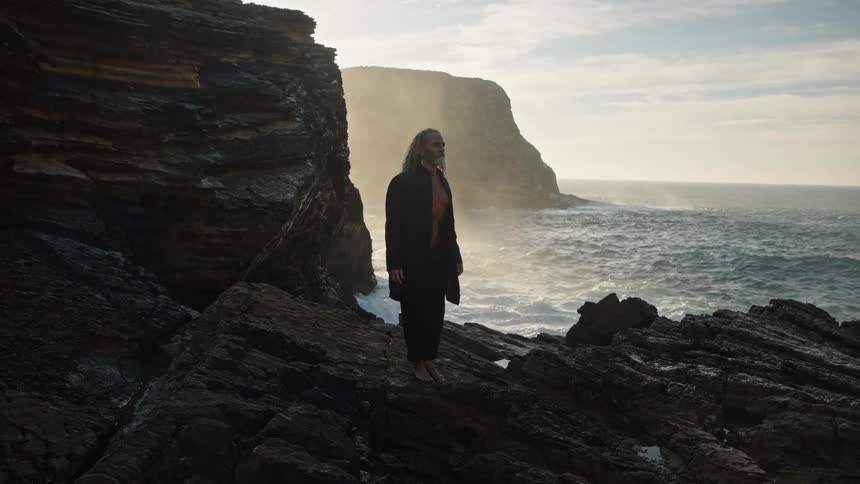 An older man with long hair standing among rocks near the sea