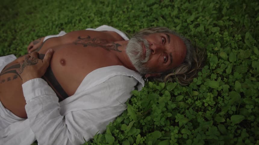 The video is a medium closeup of a smiling man with a long white beard and many tattoos who is lying down outdoors he is resting on his back in a patch of dense green clover or ivy with his eyes closed and his chest bare under an open white shi