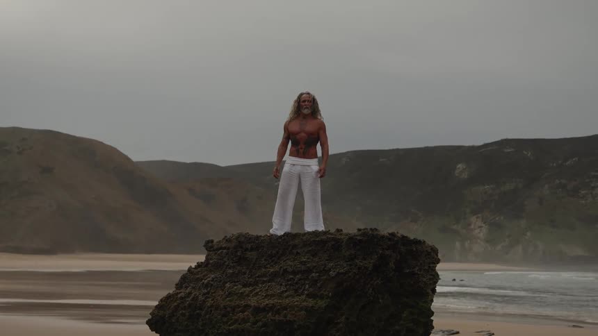 Man with long hair standing on a rock at the beach with waves in the background