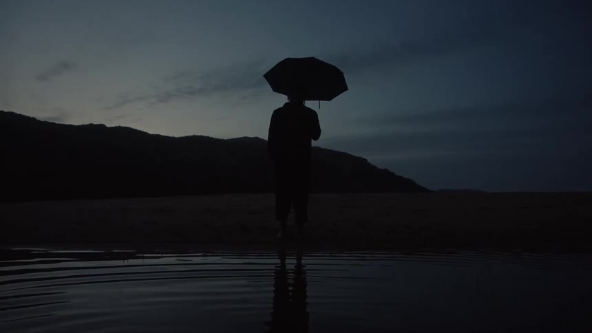 A person stands facing forward in a body of water the person holds an open umbrella above the head the figure is a dark outline against a muted sky and a line of land mass is visible on the far horizon