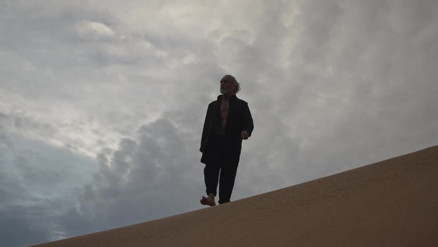 A middleaged man walks barefoot across desert sand at midday beneath a cloudy sky
