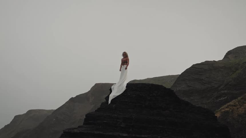A long view of a blonde bearded man wearing a light colored dress standing on a dark rocky peak against a background of steep green hills and an overcast sky
