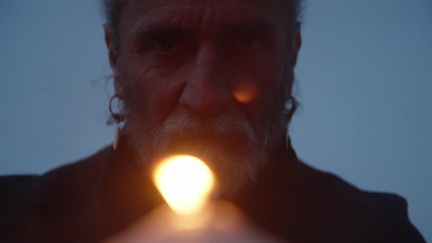 This is an extreme closeup shot of an older mans face and chest in soft bluehour lighting with a focus on his beard and a simple silver earring he is holding a single burning candle in the foreground with the warm bright flame providi