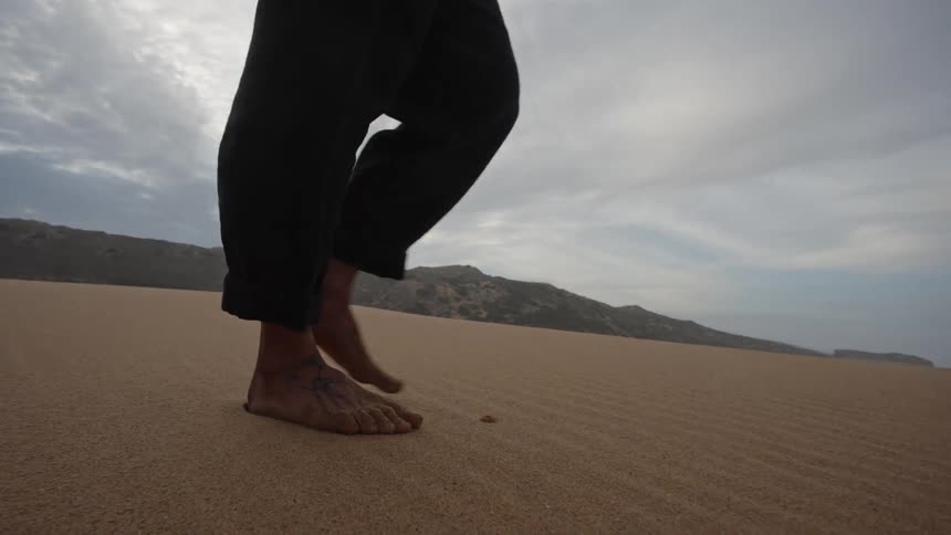 A man with long hair and a beard wearing dark clothes walking slowly barefoot across a sand dune with hills visible on the horizon under a cloudy sky