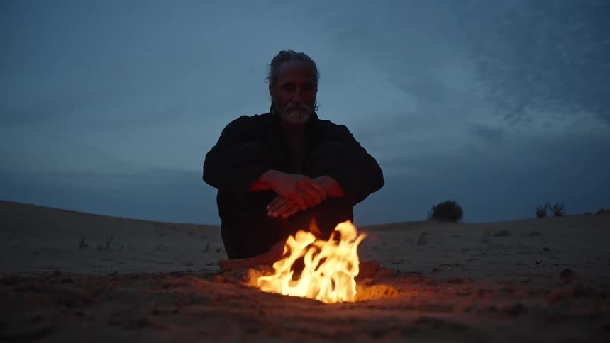 Man sits by a small campfire on the sand at night