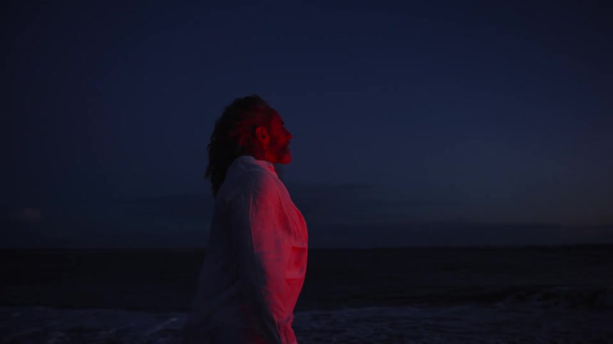 A profile medium view of a man with long hair wearing a white shirt and pants backlit by a red light on a dark beach or shoreline at twilight as small waves crash nearby
