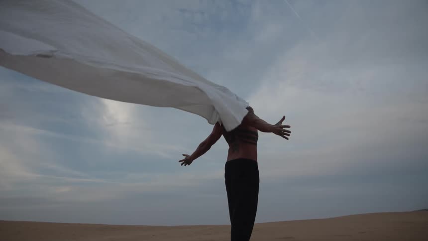 A person stands on a sandy ground with arms extended and body facing away holding a large white sheet that billows upward in the wind against a cloudy sky