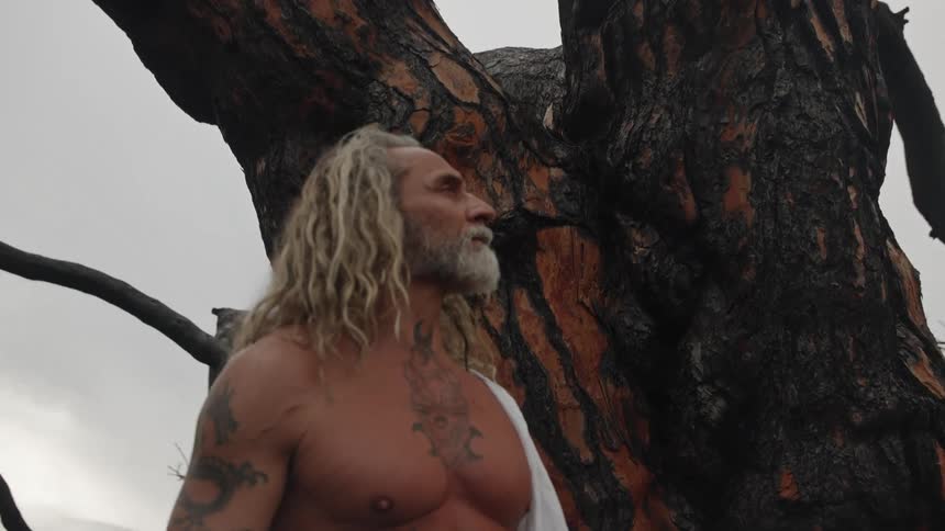 A muscular tattooed man with long blonde hair and a white shawl stands next to a burnt tree trunk in a postfire forest touching the charred bark with his hand against an overcast sky