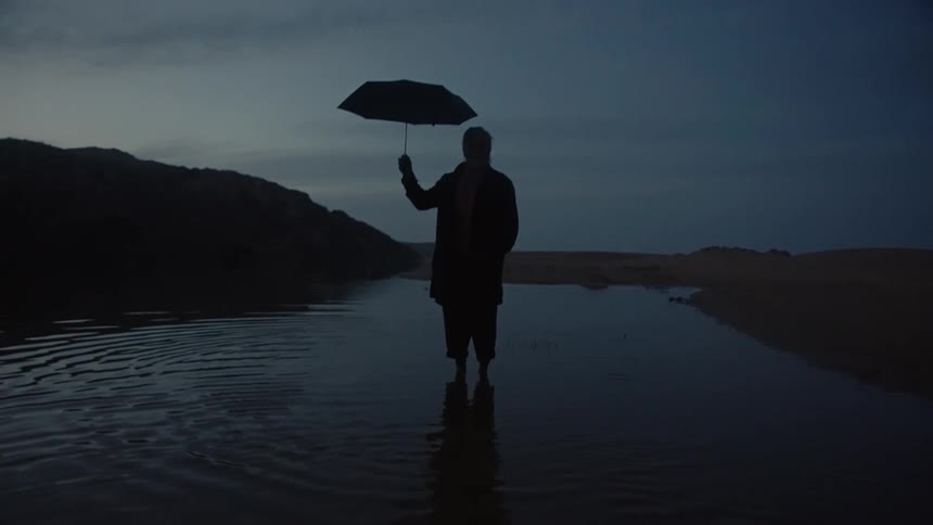 A wide low angle shot of a solitary person silhouetted against the dark twilight sky as they stand in shallow water holding an open black umbrella with wet sand and dark hills in the background