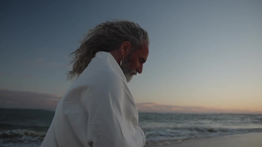 An old hippie man standing on the beach looking up and reflecting