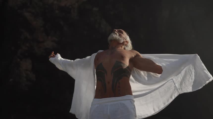 The video is a medium shot of a tattoed man with a long white beard and his hair tied in a top knot standing against a dark background he is wearing an open white shirt exposing his chest and arms which are covered in dark tattoos and he looks