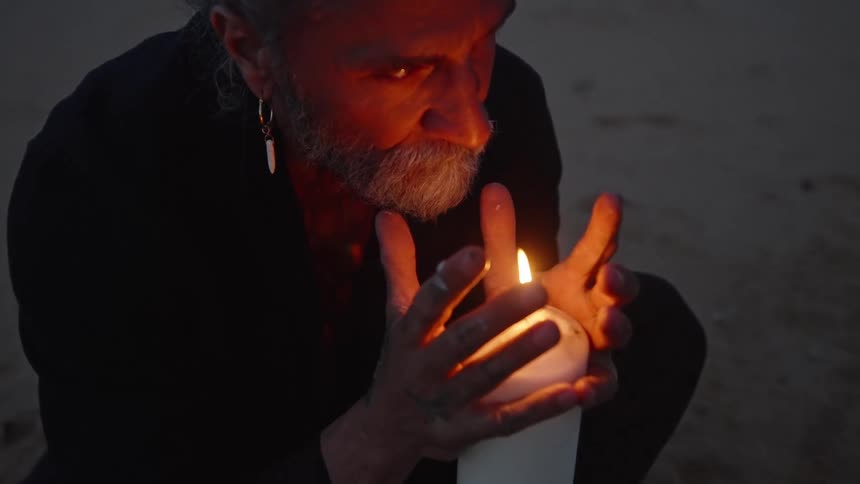 An old man crouching on the sand holding his hands around a lit candle
