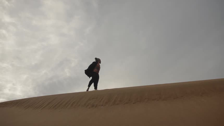 A figure walks alone across the crest of a sand dune moving from left to right as a silhouette against a pale cloudy sky