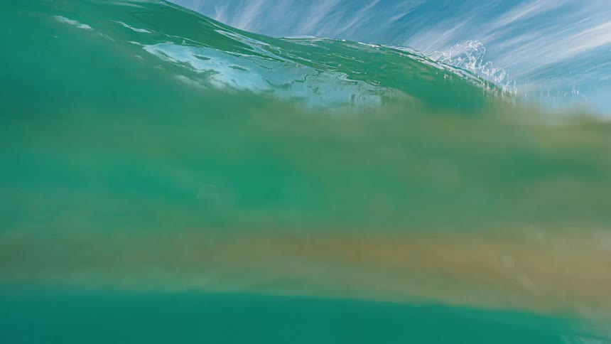 A wide view of an emerald green ocean wave crests over the camera transitioning to an underwater view of deep blue water under a bright sky with wispy clouds