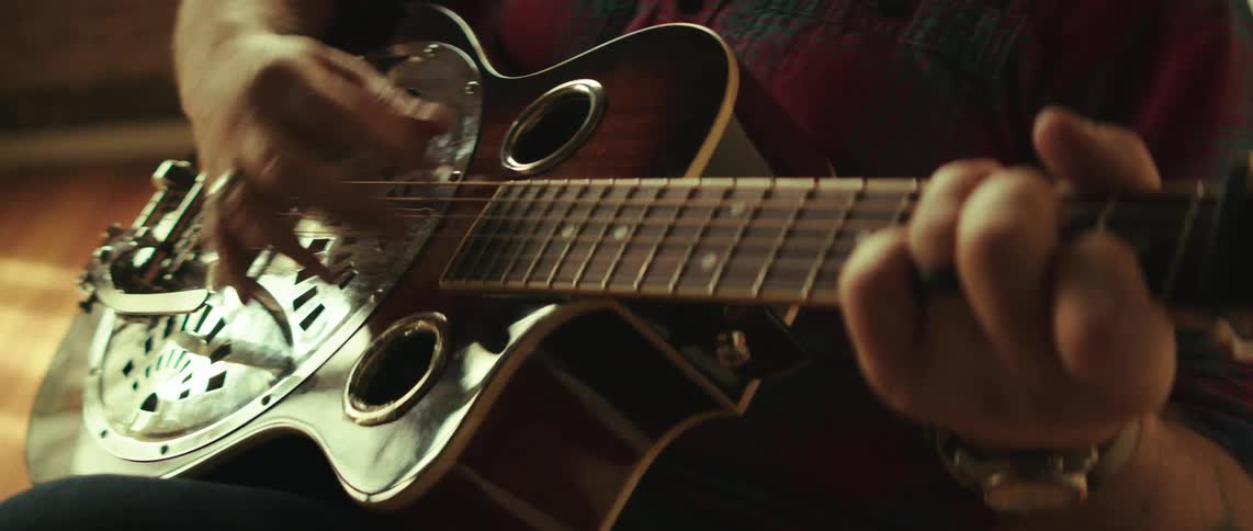 A close view of a person with rings on their fingers strumming a resonator guitar with a silver cover plate while wearing a red and black plaid shirt