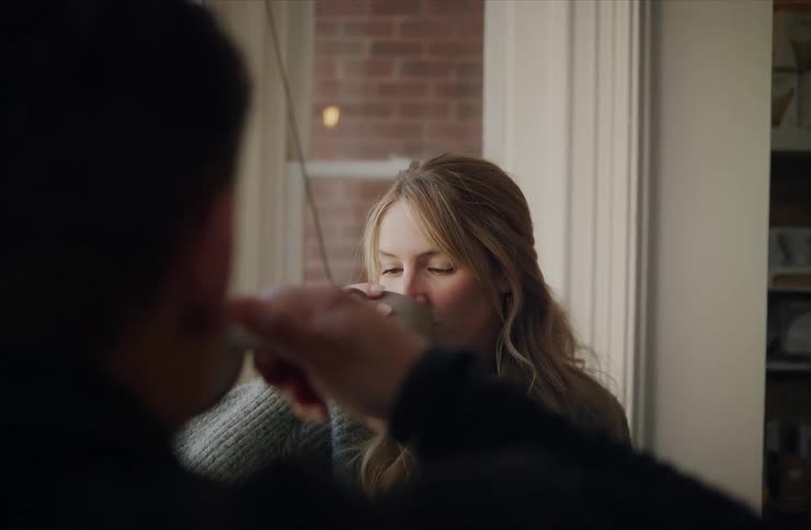 A woman with long blonde hair and a gray sweater is standing by a window taking a sip from a large rustic mug before looking up she appears to be having a conversation with a dark outoffocus figure positioned in the foreground